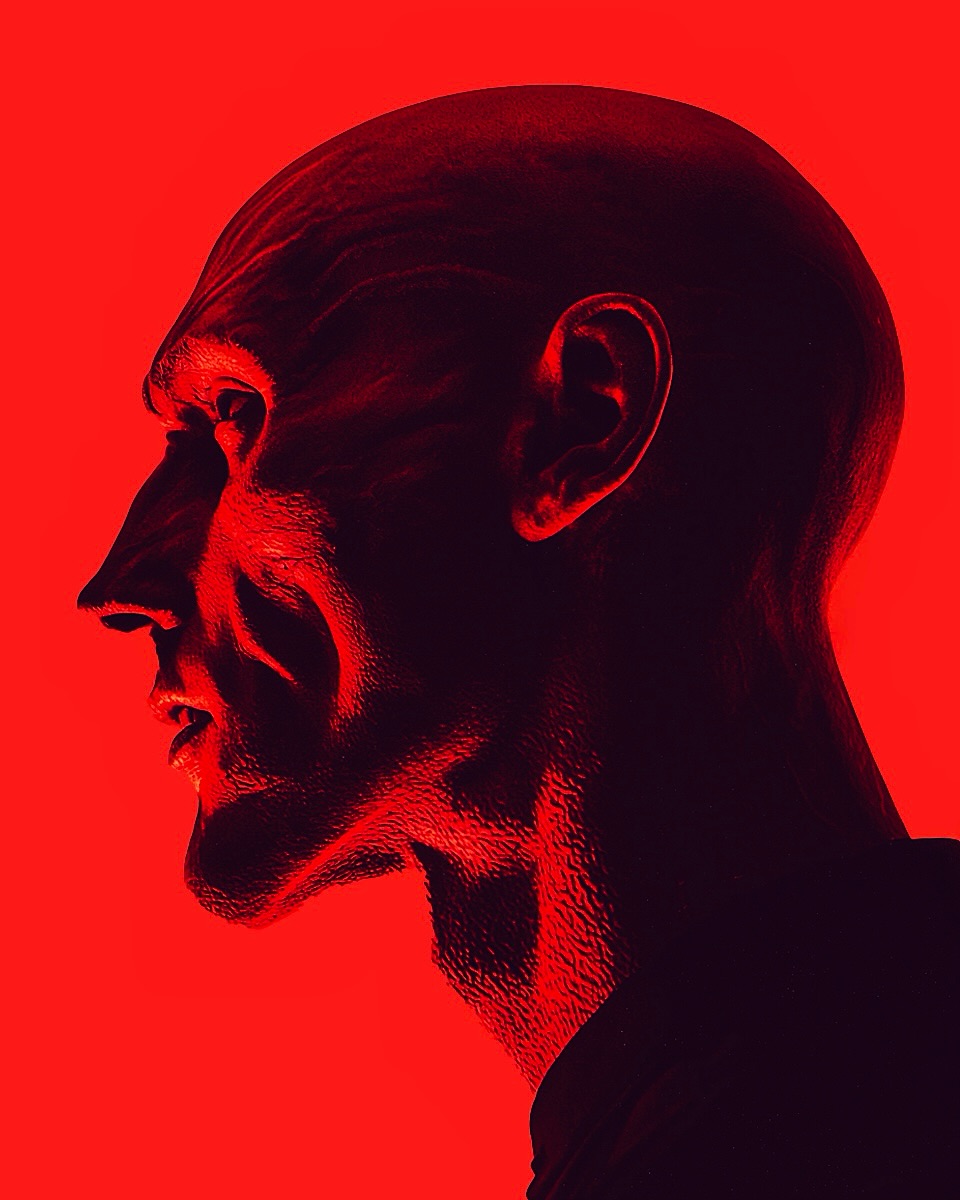 Portrait of a bald adult male in profile with red lighting. Intense facial expression and textured skin are visible against a red background.