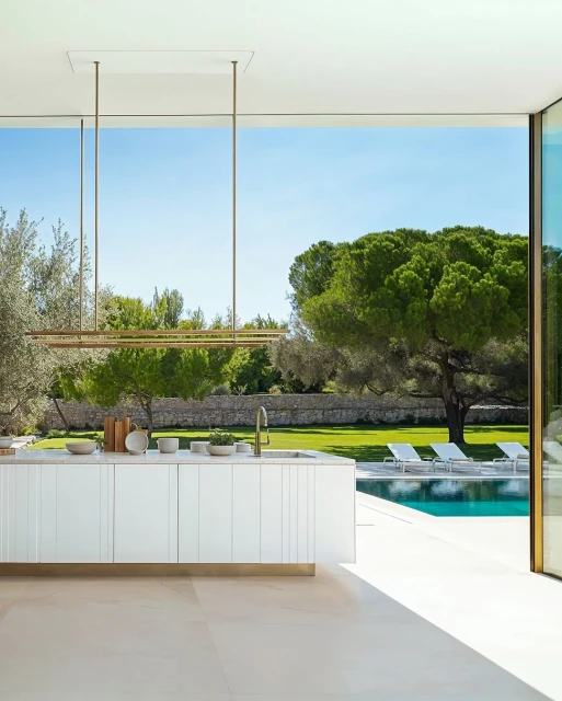 Bright, modern kitchen island overlooks a pool and green lawn with trees. White cabinetry and stone countertop. Luxury living concept.