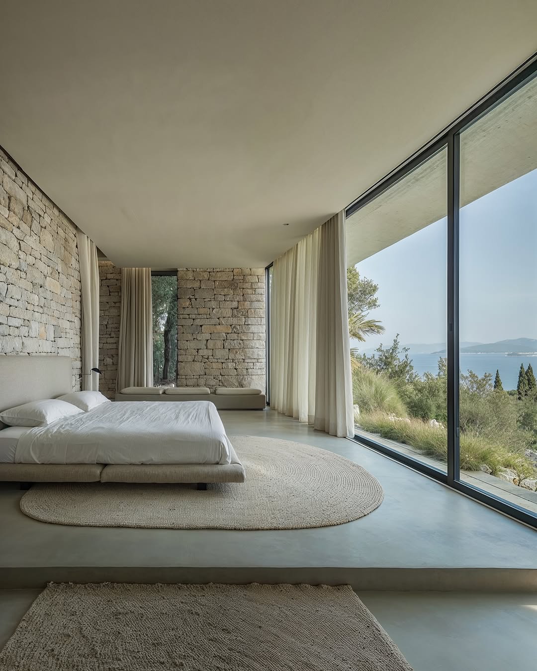 Bedroom with stone walls, modern bed, and large windows offering a view of the sea and lush greenery, creating a serene interior.