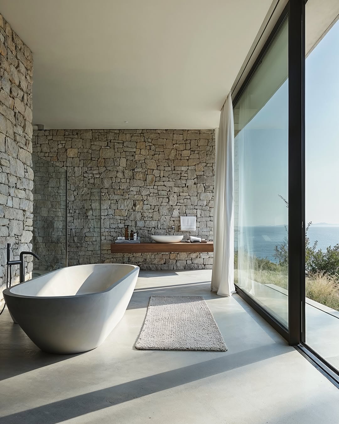 Modern bathroom with stone wall, freestanding tub, wood vanity, large windows with ocean view and neutral colors for spa-like retreat.