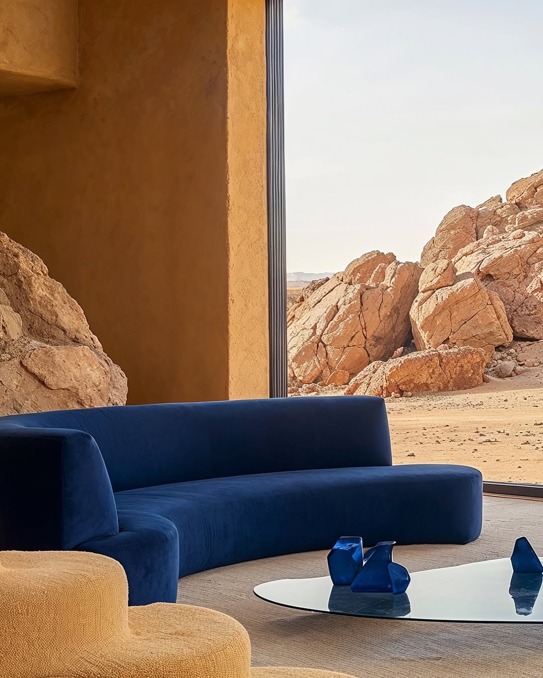 Interior of a living room featuring a curved blue sofa, glass coffee table, and desert landscape visible through a large window.