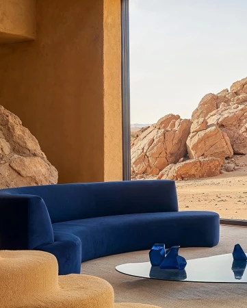 Interior of a living room featuring a curved blue sofa, glass coffee table, and desert landscape visible through a large window.