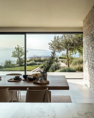 Bright and airy dining room interior with a wooden table set for a meal, opening to a scenic outdoor view of the sea and garden.
