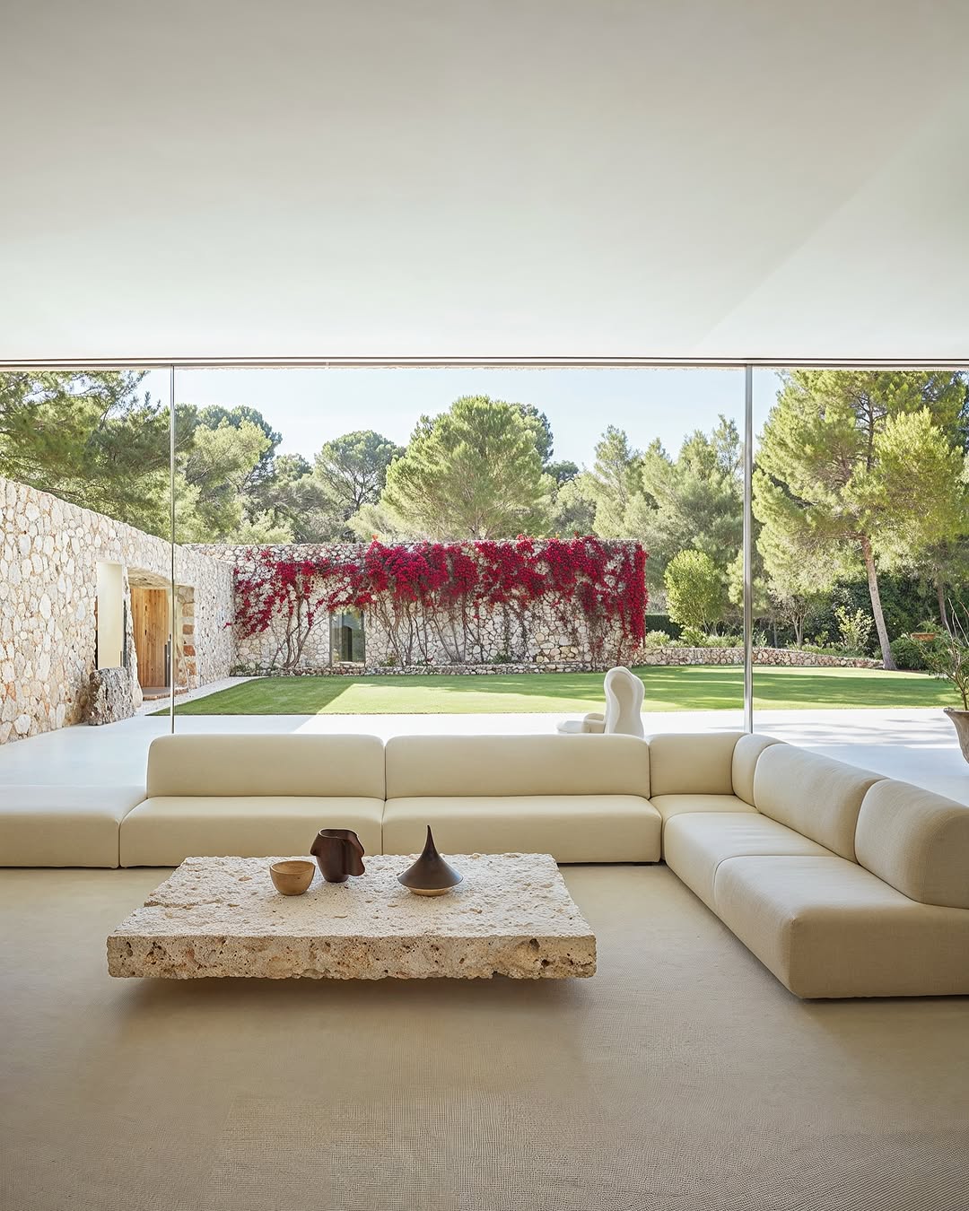 Living room interior with a large sectional sofa, a travertine coffee table, and floor-to-ceiling windows overlooking a garden.