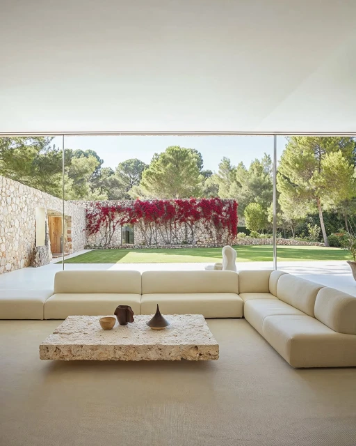 Living room interior with a large sectional sofa, a travertine coffee table, and floor-to-ceiling windows overlooking a garden.