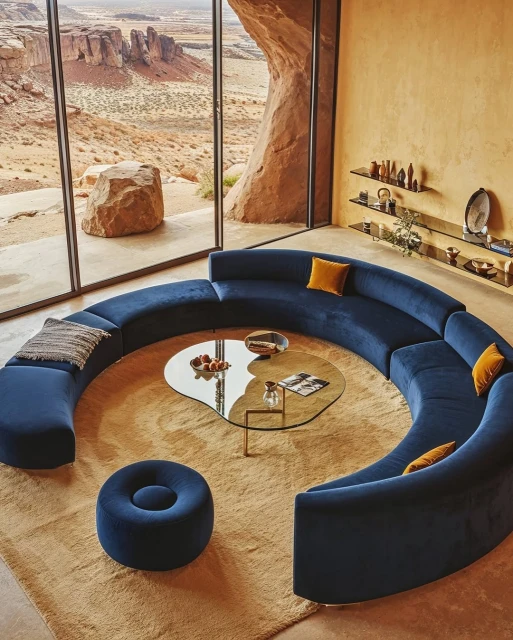 Living room interior with a curved blue sofa, round glass coffee table, ottoman, and desert landscape seen through a large window.