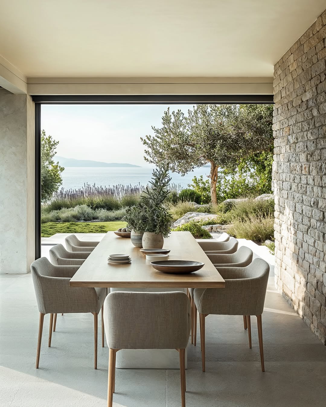Outdoor dining area with a long wooden table, upholstered chairs, and view of a lake and trees, creating a serene atmosphere.