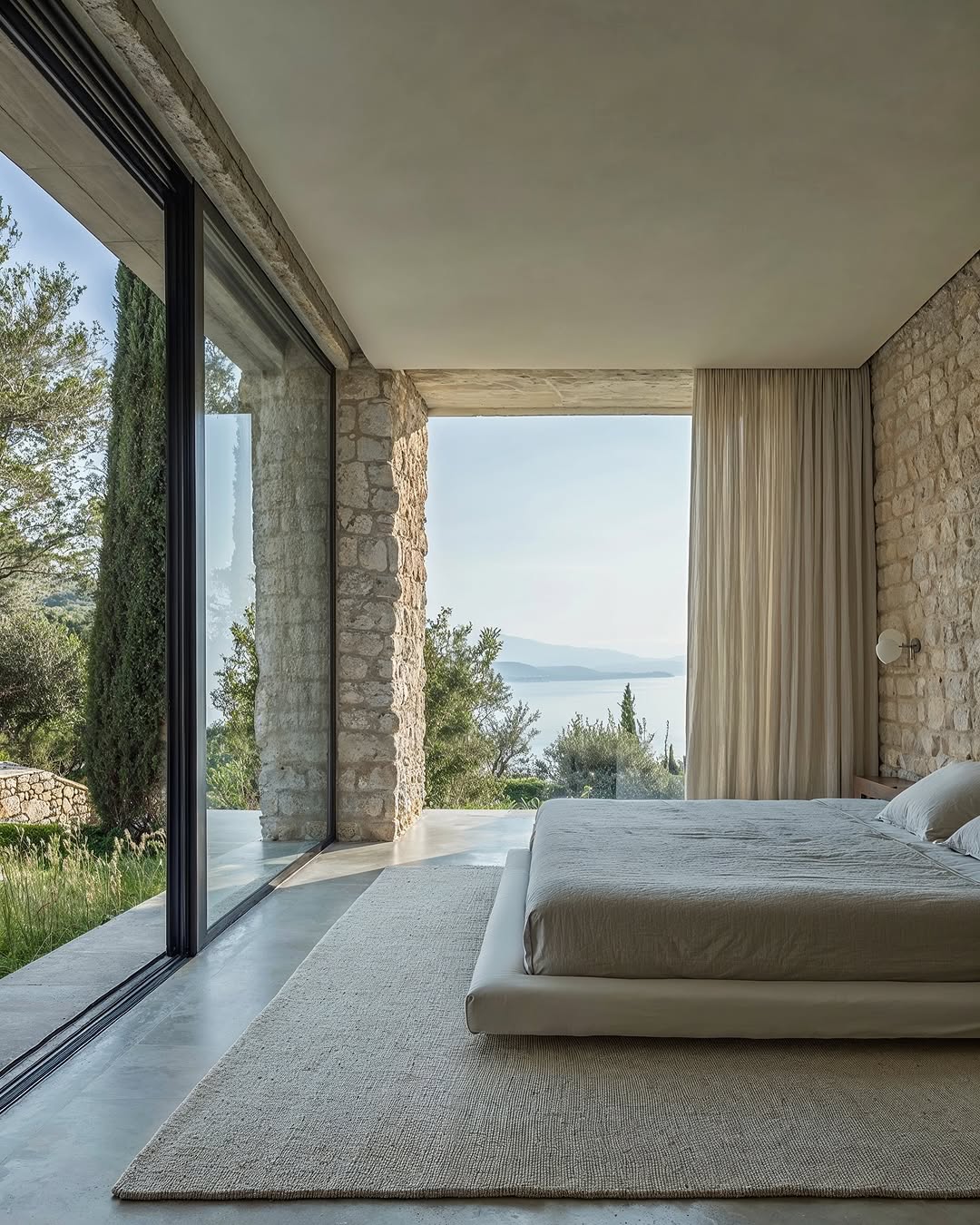Minimalist bedroom featuring a large platform bed, neutral-toned rug, and floor-to-ceiling windows with a view of greenery and the sea.