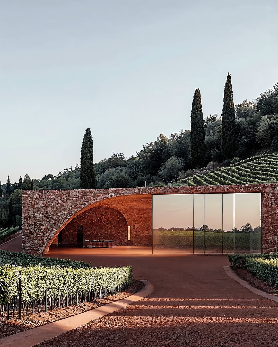 Exterior of modern red brick winery building with arched entryway and glass wall, surrounded by vineyard rows and green hillside.