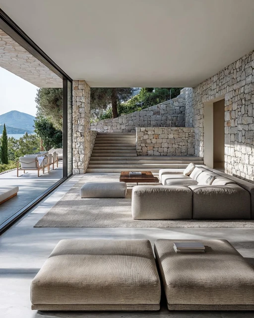 Living room with modular sofa and stone walls, featuring open glass doors to a terrace with outdoor furniture and a sea view.