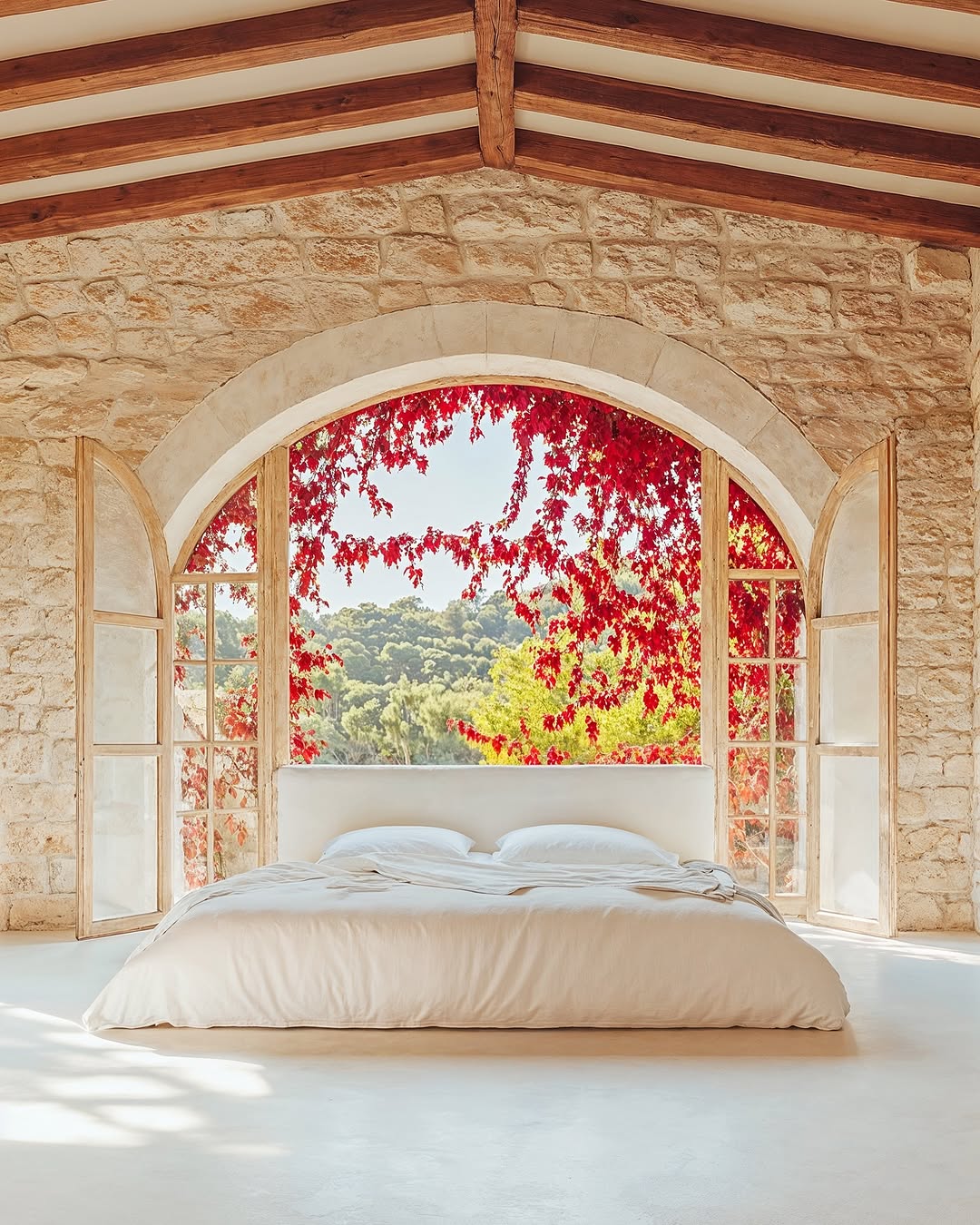 A bed is framed by an arched window with red vines, inside a stone room with a wooden ceiling in a naturally lit interior.