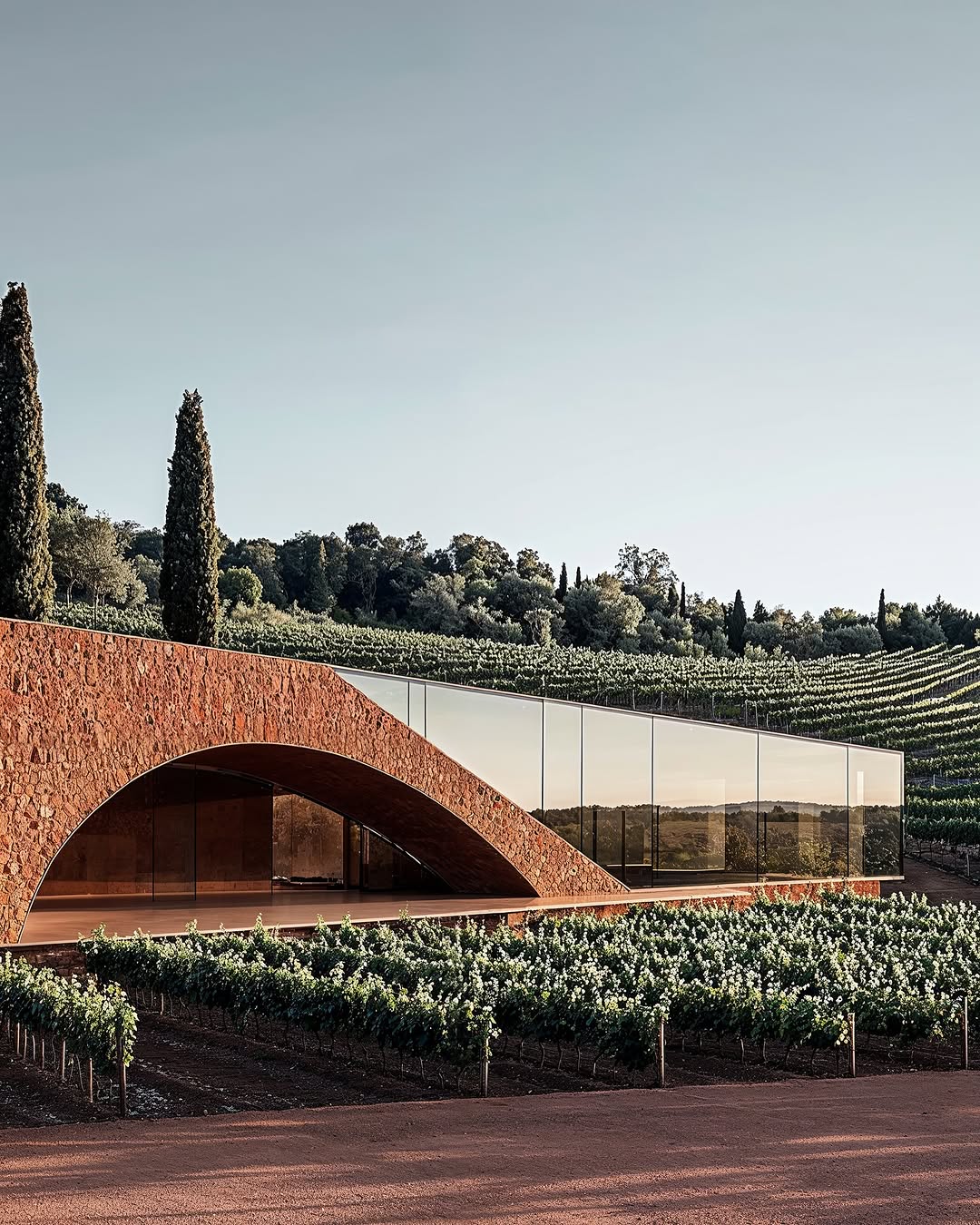 Modern architectural building with brick archway entrance, glass walls, and vineyard in rural landscape setting under a clear sky.