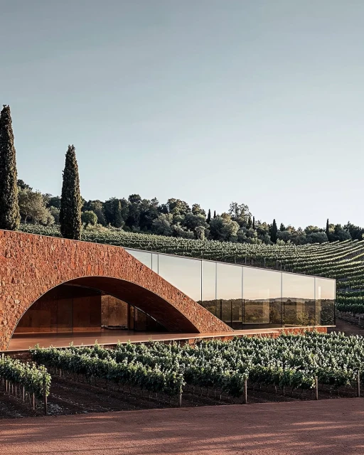 Modern architectural building with brick archway entrance, glass walls, and vineyard in rural landscape setting under a clear sky.