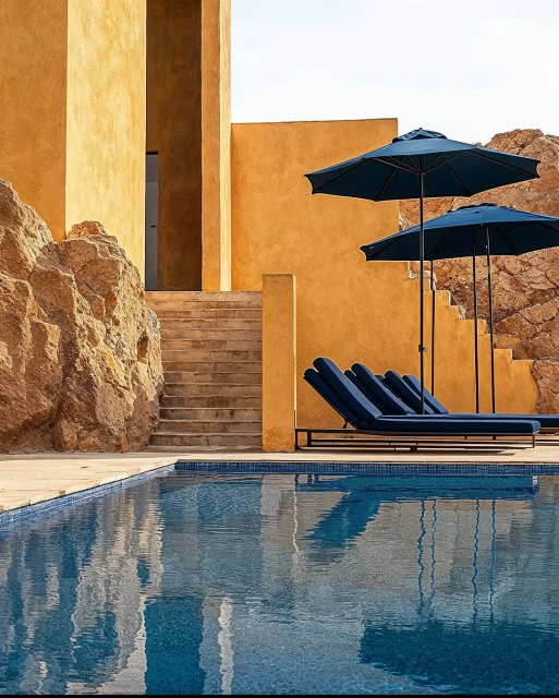 Swimming pool with lounge chairs and sun umbrellas next to a tan building and rock formation on a sunny day.