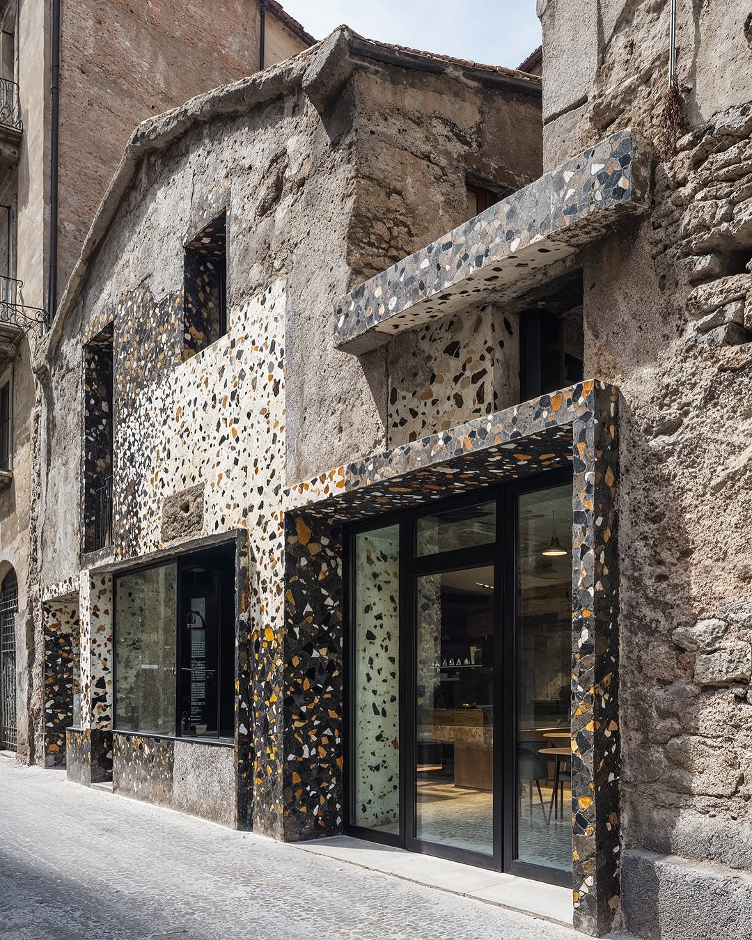 Exterior of a modern building with a terrazzo facade in a narrow street, featuring large windows and an inviting entrance.