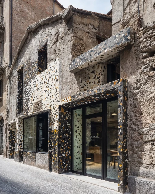 Exterior of a modern building with a terrazzo facade in a narrow street, featuring large windows and an inviting entrance.