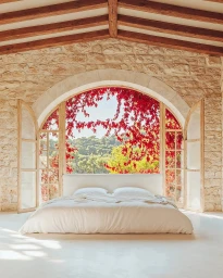 A low-slung white bed centered in front of a large arched window framing red flowering vines against a stone wall interior.