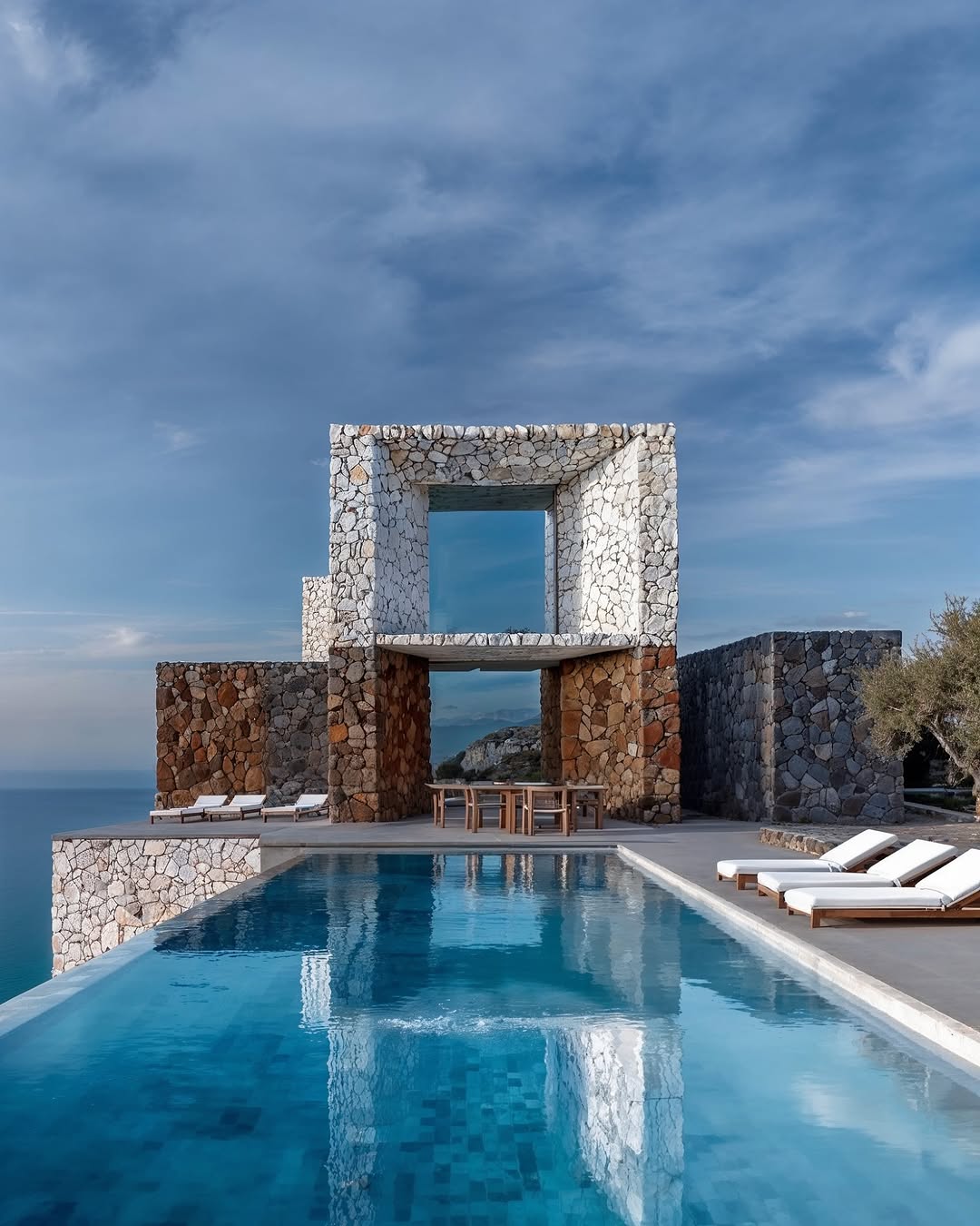 Modern stone house with infinity pool overlooking the ocean on a sunny day, architectural design showcases luxury coastal living.