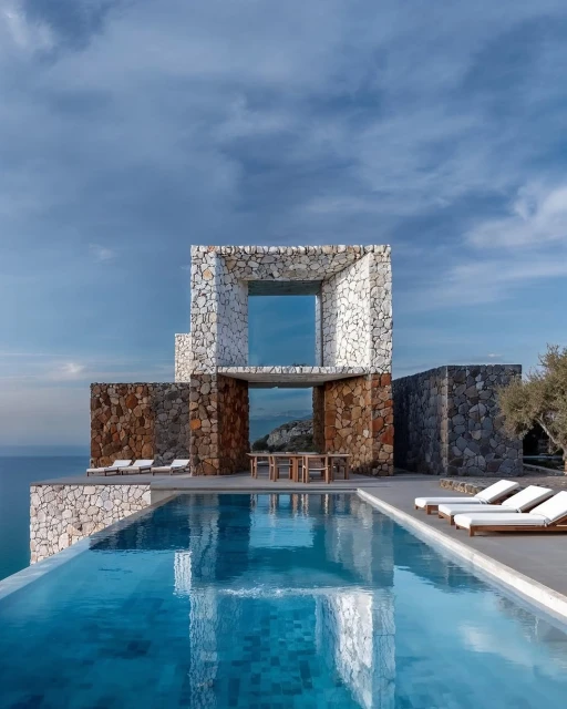 Modern stone house with infinity pool overlooking the ocean on a sunny day, architectural design showcases luxury coastal living.