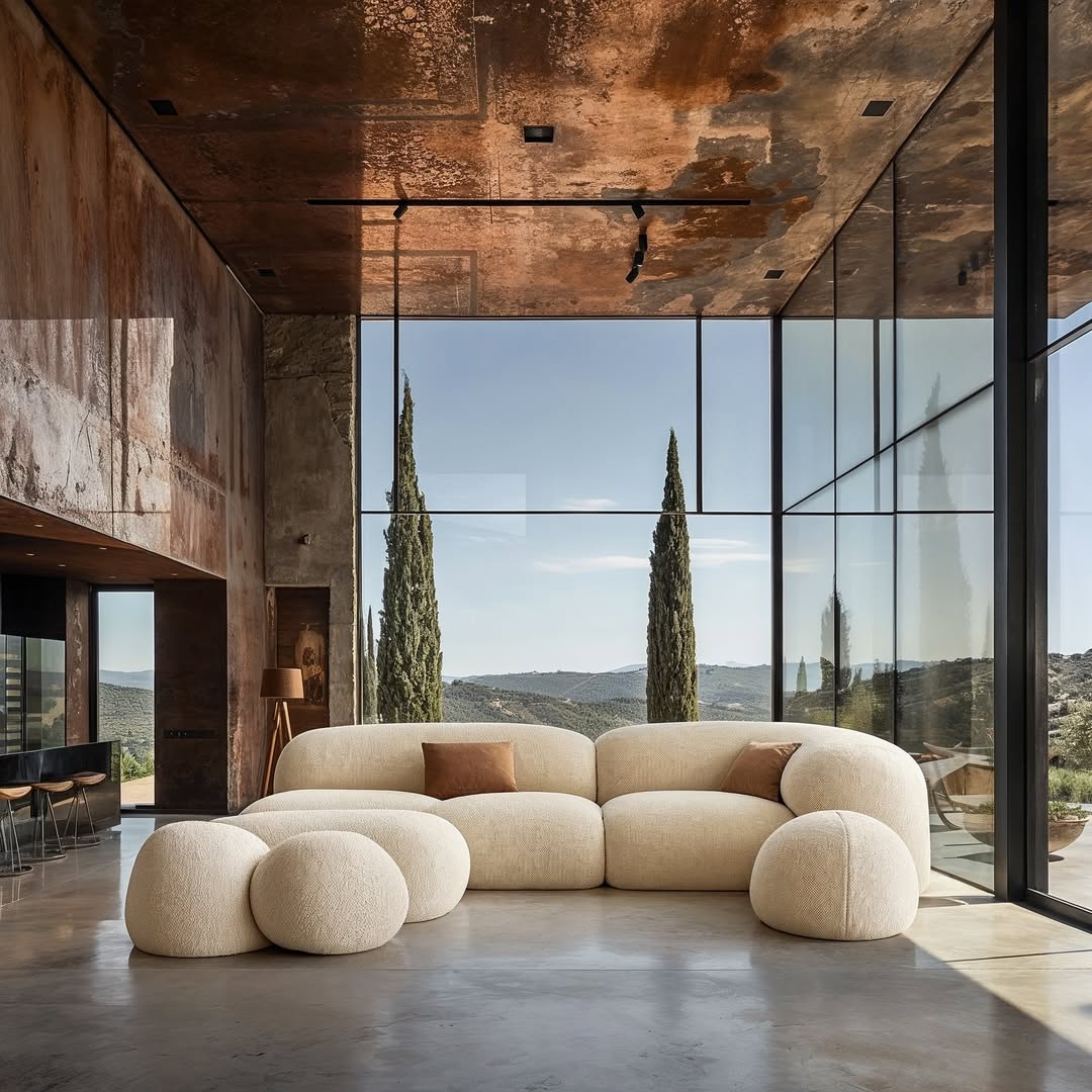 Interior view of a modern living room with a modular sofa, large windows, and a view of the landscape. Rustic walls and neutral tones.