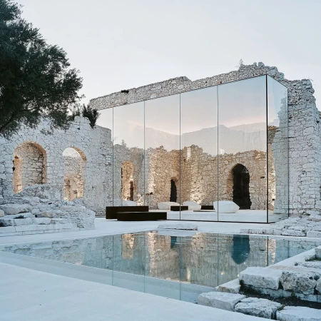 Modern architecture meets ancient ruins, featuring a glass-walled structure reflecting stone walls and a pool on a bright day.