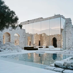 Modern glass building facade inserted within historic stone ruins beside a reflecting pool under an overcast sky.