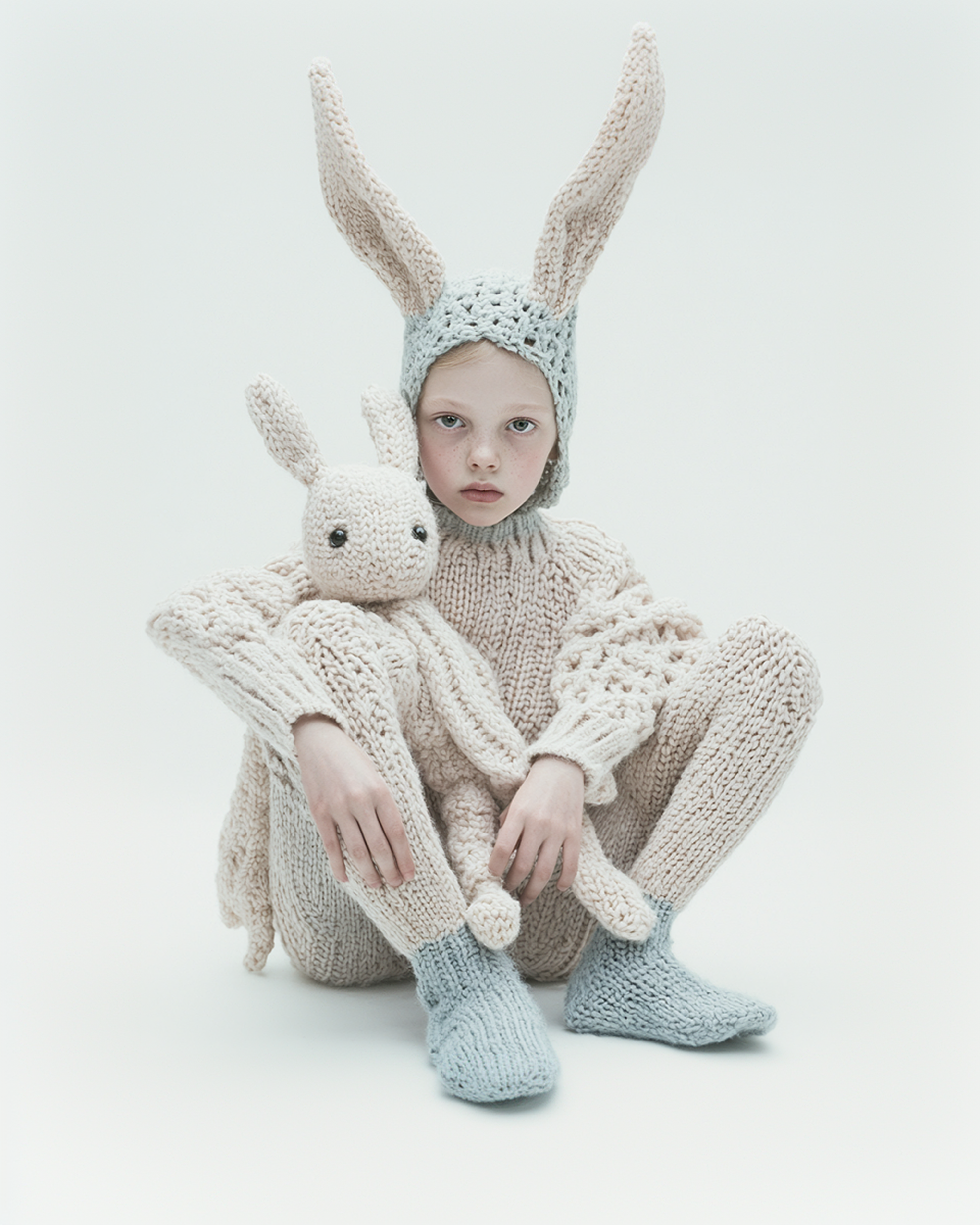Child dressed in a knitted bunny costume with a matching stuffed bunny toy, sitting against a white backdrop, looking directly at the camera.