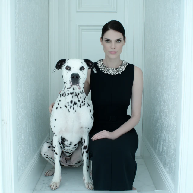 Elegant woman in a black dress poses with a Dalmatian dog in a white hallway, creating a striking contrast and sophisticated ambiance.