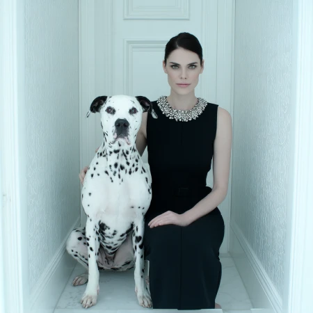 Elegant woman in a black dress poses with a Dalmatian dog in a white hallway, creating a striking contrast and sophisticated ambiance.