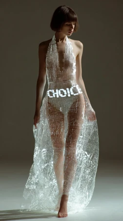 Woman in a halter dress with 'CHOICE' text design, standing barefoot in a studio. The dress has a sheer, shimmering fabric.