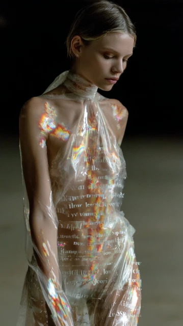 Woman poses in a transparent, text-covered dress against a dark backdrop, her expression serene and the lighting accentuating her features.