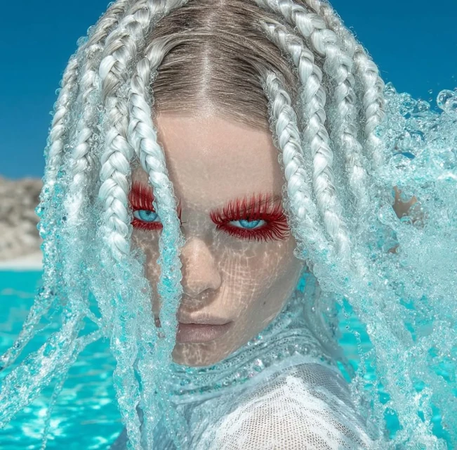 Close-up portrait of a woman in water, featuring braided white hair, red eyelashes, and bright blue eyes under clear blue sky.