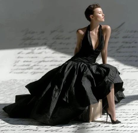 Elegant woman in a black halter dress sitting on a block in a light studio with script lettering in the backdrop. Fashion editorial.