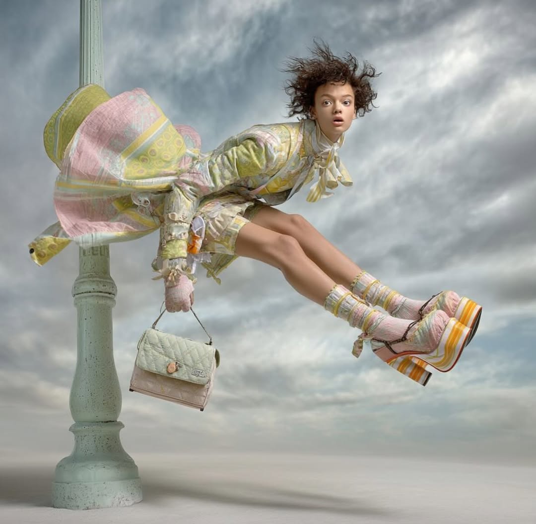 A young woman in a patterned pastel-colored outfit and platform shoes sits atop a light pole against a cloudy sky holding a purse.