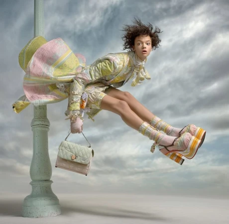 A young woman in a patterned pastel-colored outfit and platform shoes sits atop a light pole against a cloudy sky holding a purse.