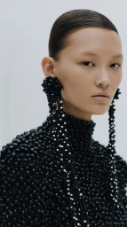 Close-up portrait of a woman wearing an intricate black beaded high-neck garment and long beaded earrings against a plain white background.
