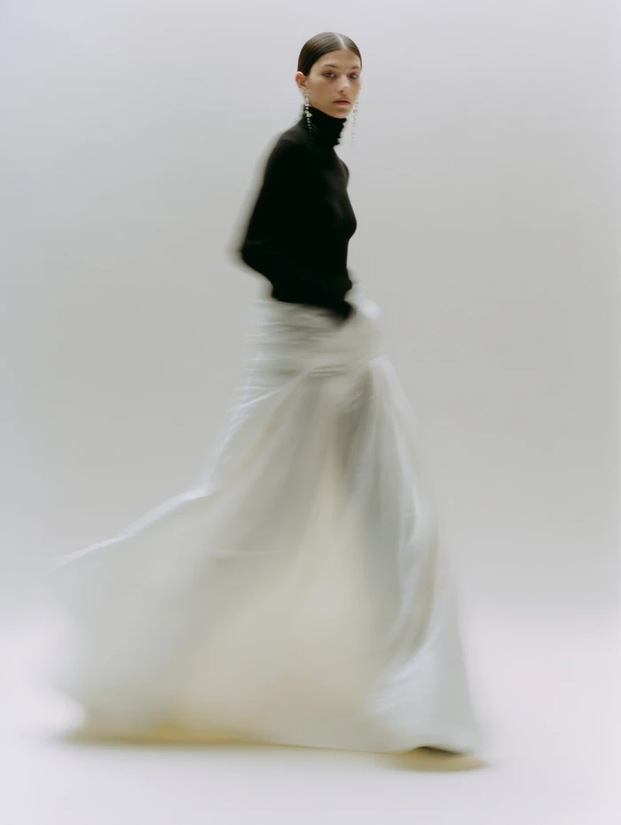 Model in black turtleneck and flowing white skirt with motion blur against a neutral studio background