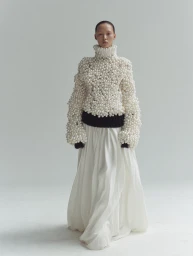 Model wearing a heavily textured turtleneck sweater covered in white pearls and a long white skirt against a plain studio background.