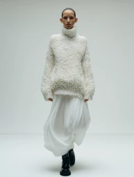Model wearing a heavily textured white knit turtleneck sweater and flowing skirt against a plain white background