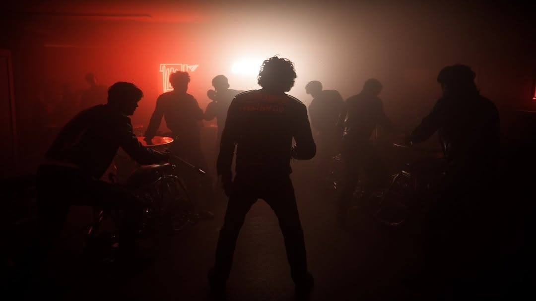 Silhouetted group of people in a dimly lit indoor space with smoke, lit by red and white lights, some on motorcycles.