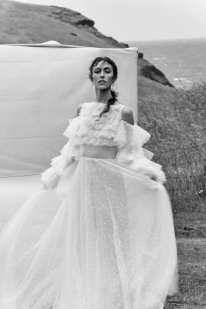 Black and white portrait of a woman wearing an elaborate ruffled white gown standing outdoors near a coastal hill by Rupert Laycock