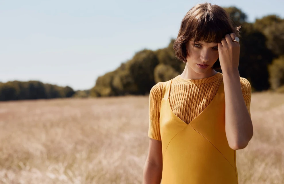 Brunette woman in yellow dress poses in sunlit field with trees in background. Fashion, portrait, outdoor, style, nature, beauty concept.