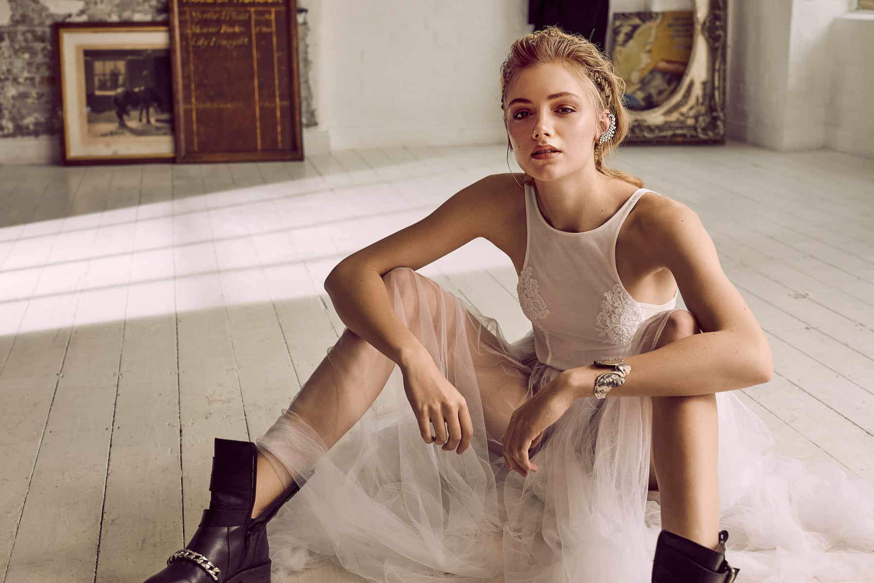 Woman in a white dress and black boots sits on a white wood floor in a sunlit room with artwork and a vintage sign on the wall.