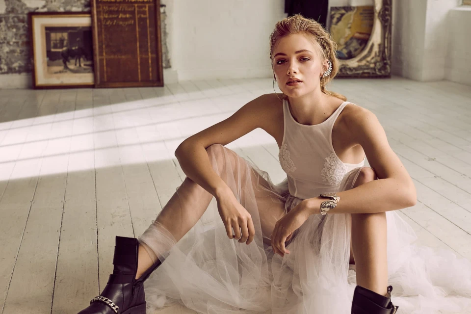 Woman in a white dress and black boots sits on a white wood floor in a sunlit room with artwork and a vintage sign on the wall.