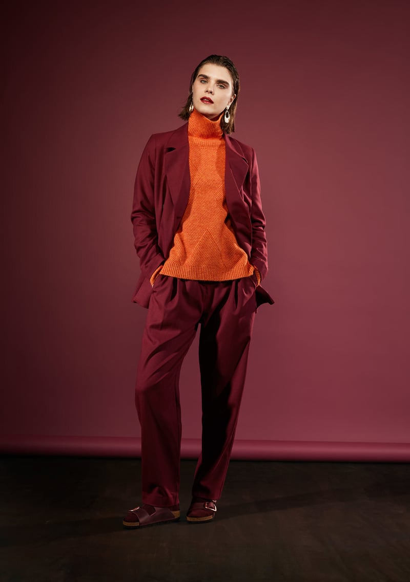 Woman in a maroon suit and orange turtleneck poses in a studio against a plum backdrop, casting a confident and stylish figure.