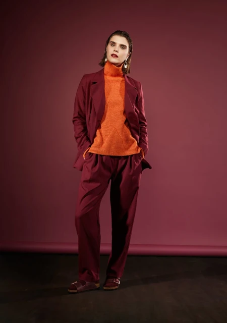 Woman in a maroon suit and orange turtleneck poses in a studio against a plum backdrop, casting a confident and stylish figure.