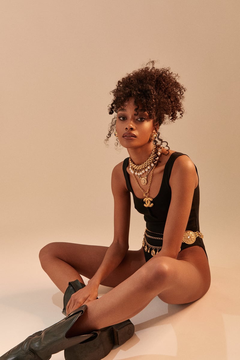 Fashion shot of a woman in a black bodysuit, cowboy boots, and gold jewelry sitting in a photo studio with a light beige background.
