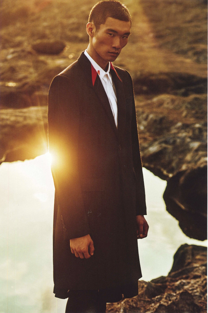 Asian male model in a dark coat with a red-trimmed collar stands against a dramatic, rocky landscape bathed in golden sunlight.