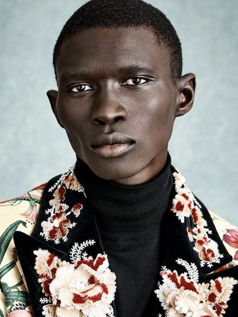Close-up portrait of Black male model in floral jacket and black turtleneck, against a muted grey backdrop, fashion photography.