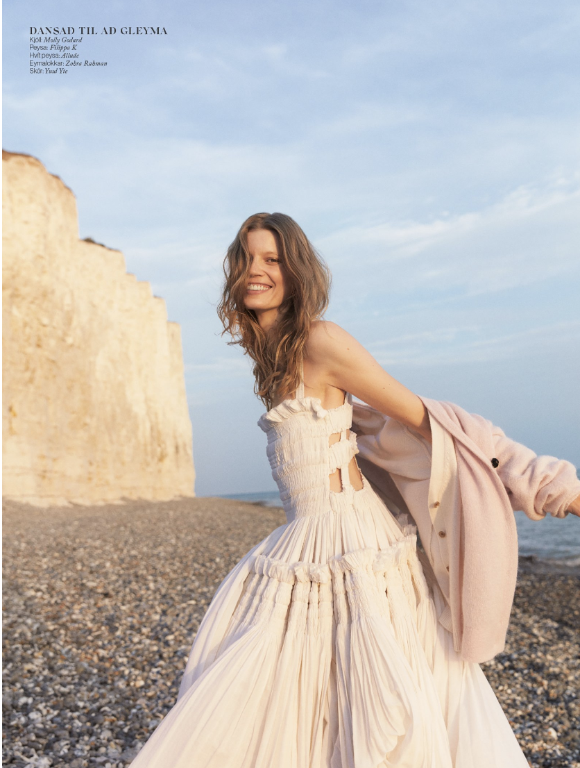 Fashion shot of a woman in a white dress and pink jacket on a pebble beach against a cliff and blue sky backdrop, smiling at the camera.